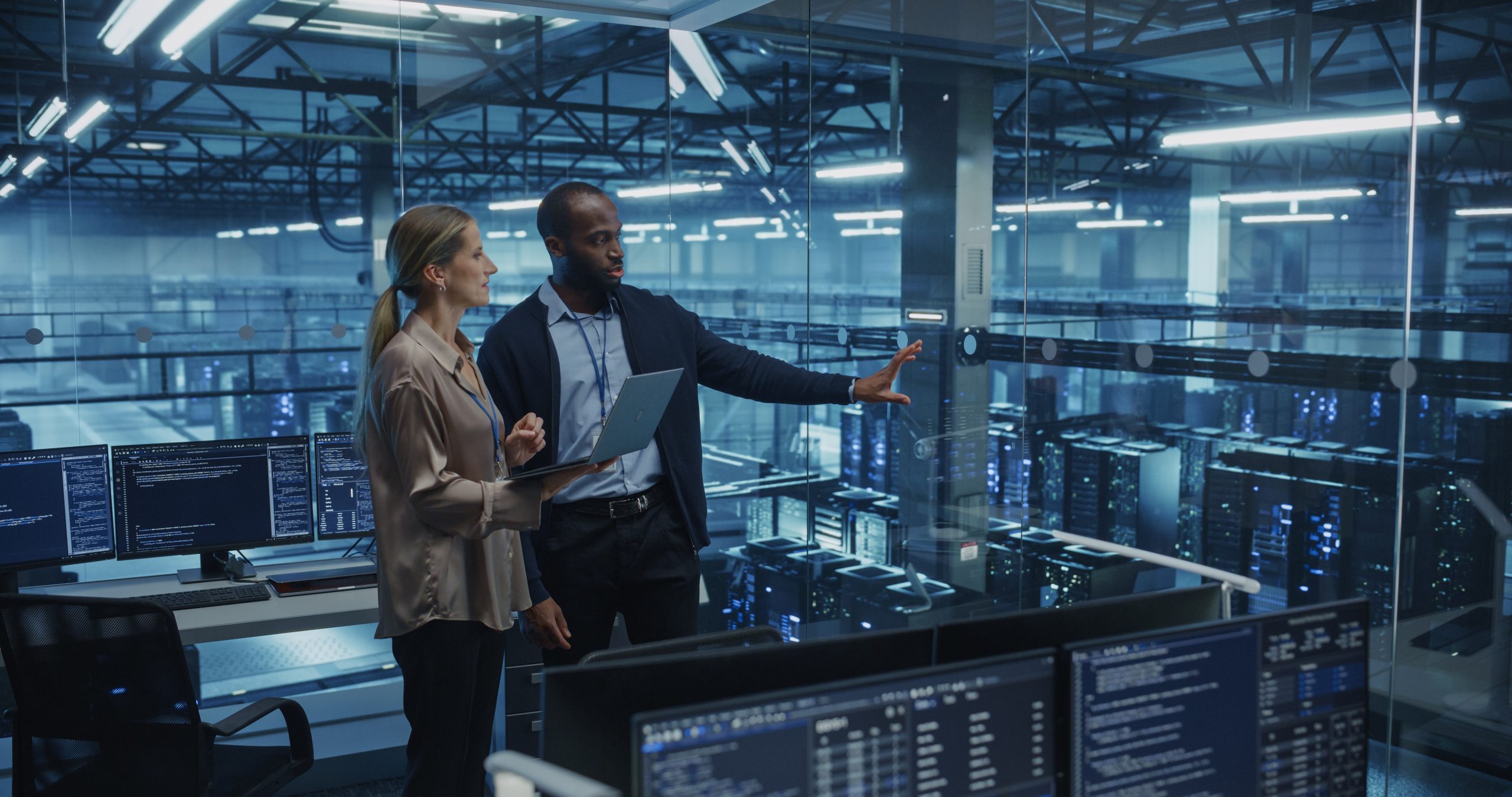 Two people with a laptop overlooking server racks, with one gesturing across the room.
