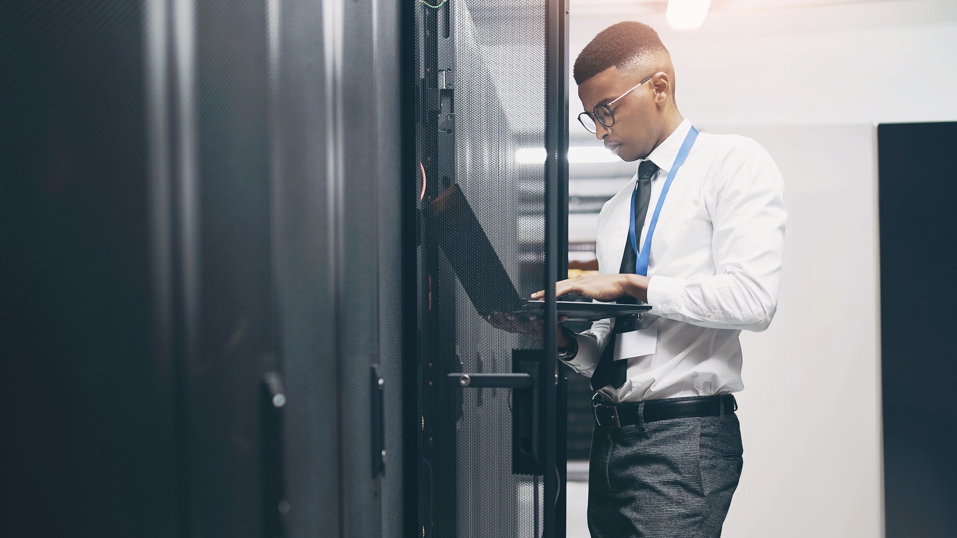 Person in a white shirt using a laptop beside equipment racks.