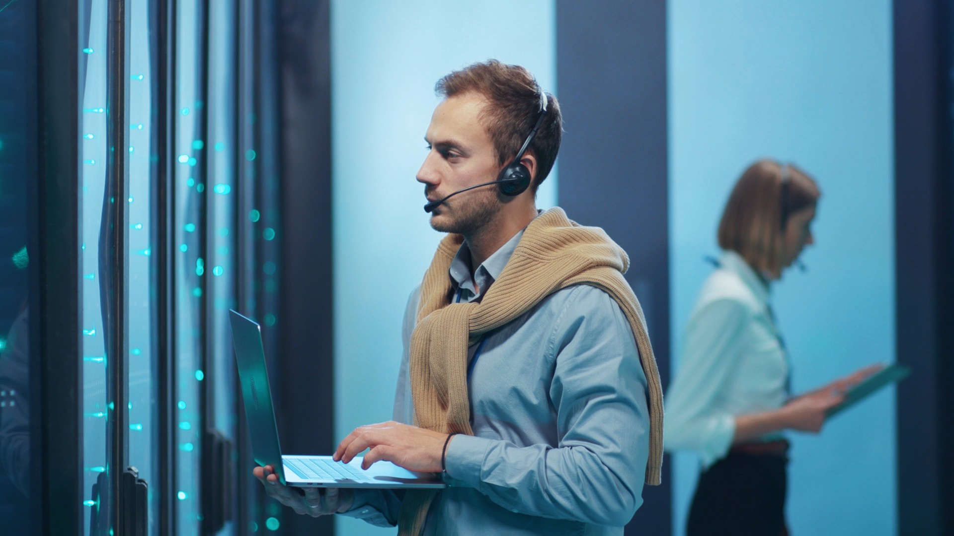 Person wearing a headset and holding a laptop beside server racks.