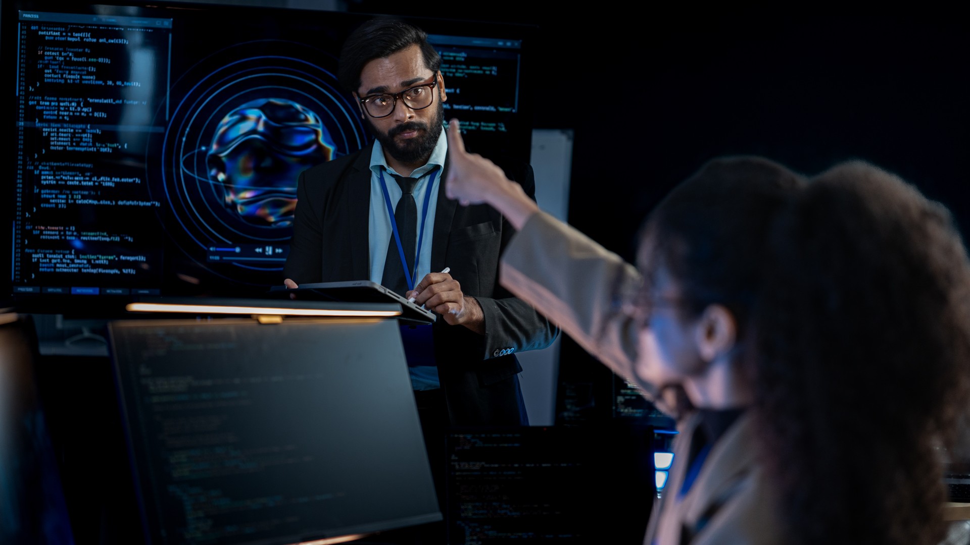 People discussing computer displays in a dark room, with one pointing toward a screen.