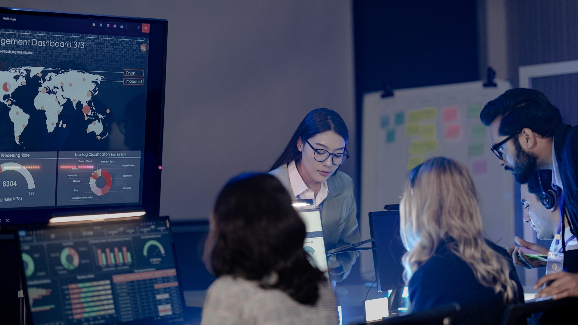 Group gathered at a workstation beside large dashboard displays with maps and charts.
