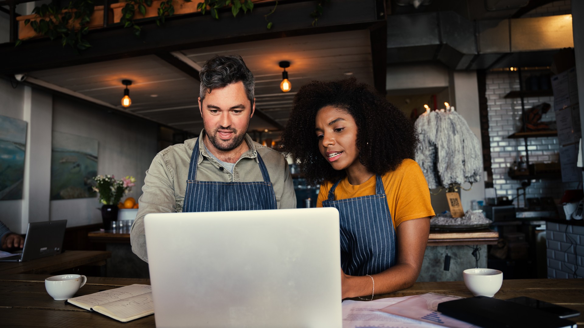 Two people wearing aprons looking at a laptop on a counter.