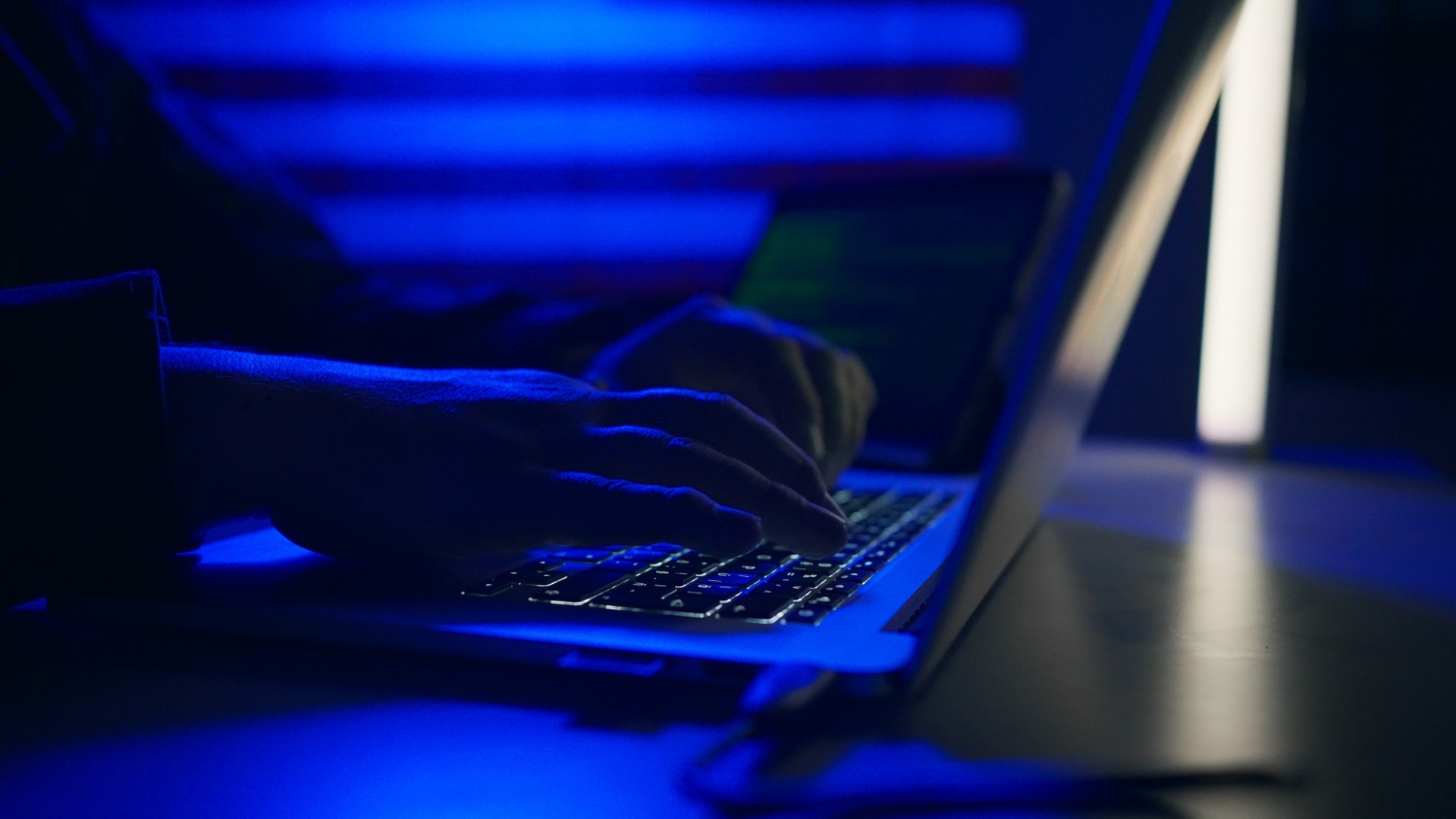 Hands typing on a laptop keyboard in a dark room lit blue.