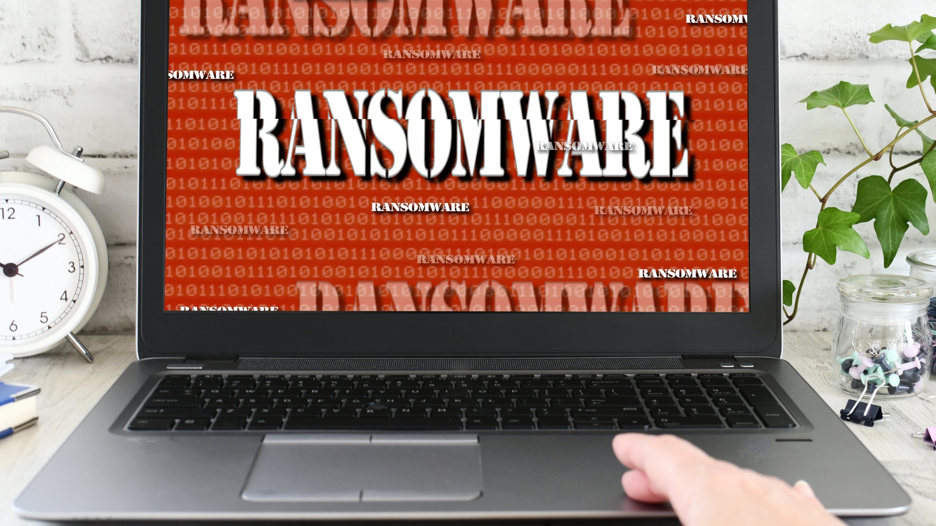 Laptop screen displaying "RANSOMWARE" in large white letters over a red binary pattern.