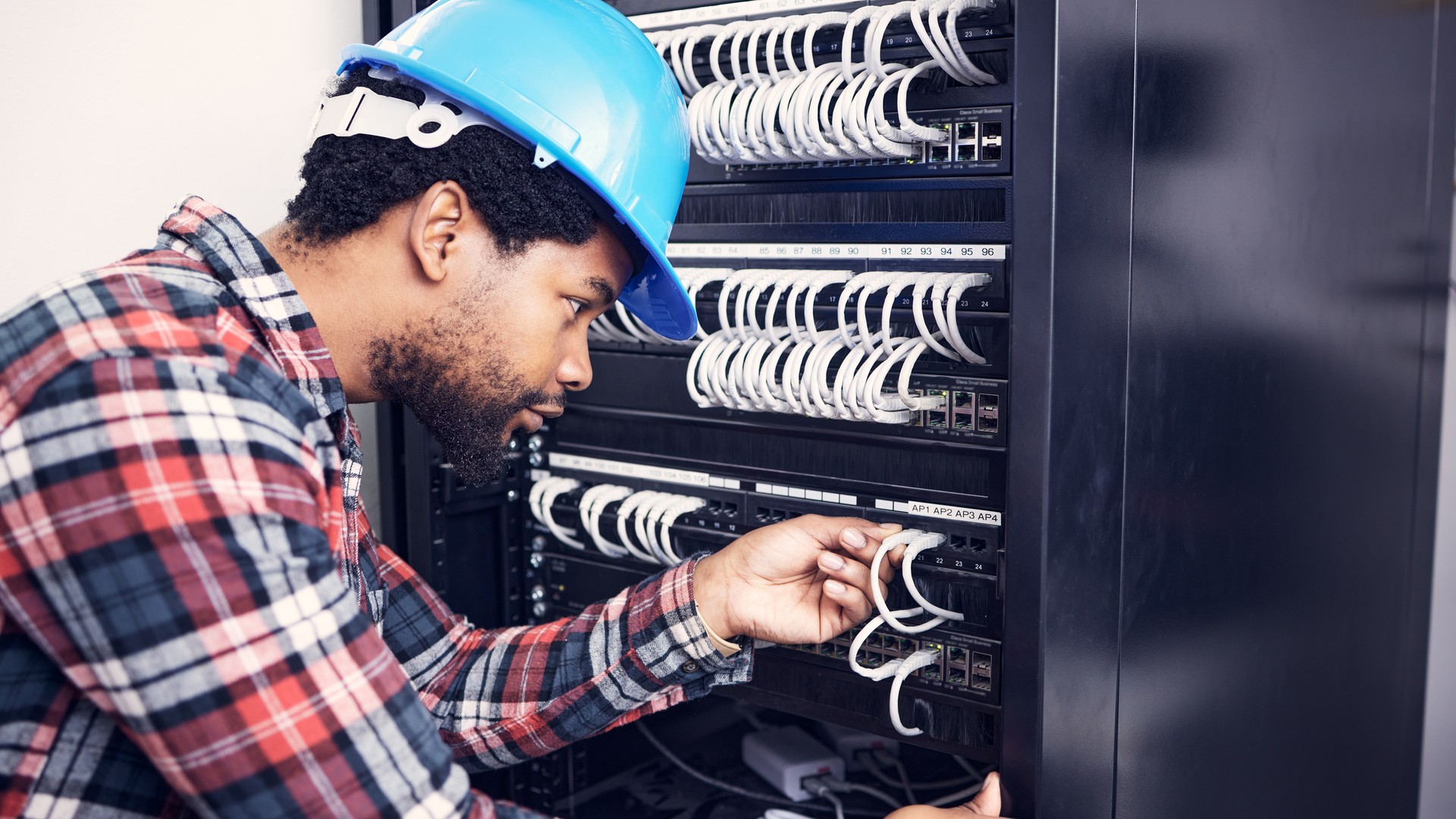 Man in a blue hard hat handling white patch cables in an open network rack.