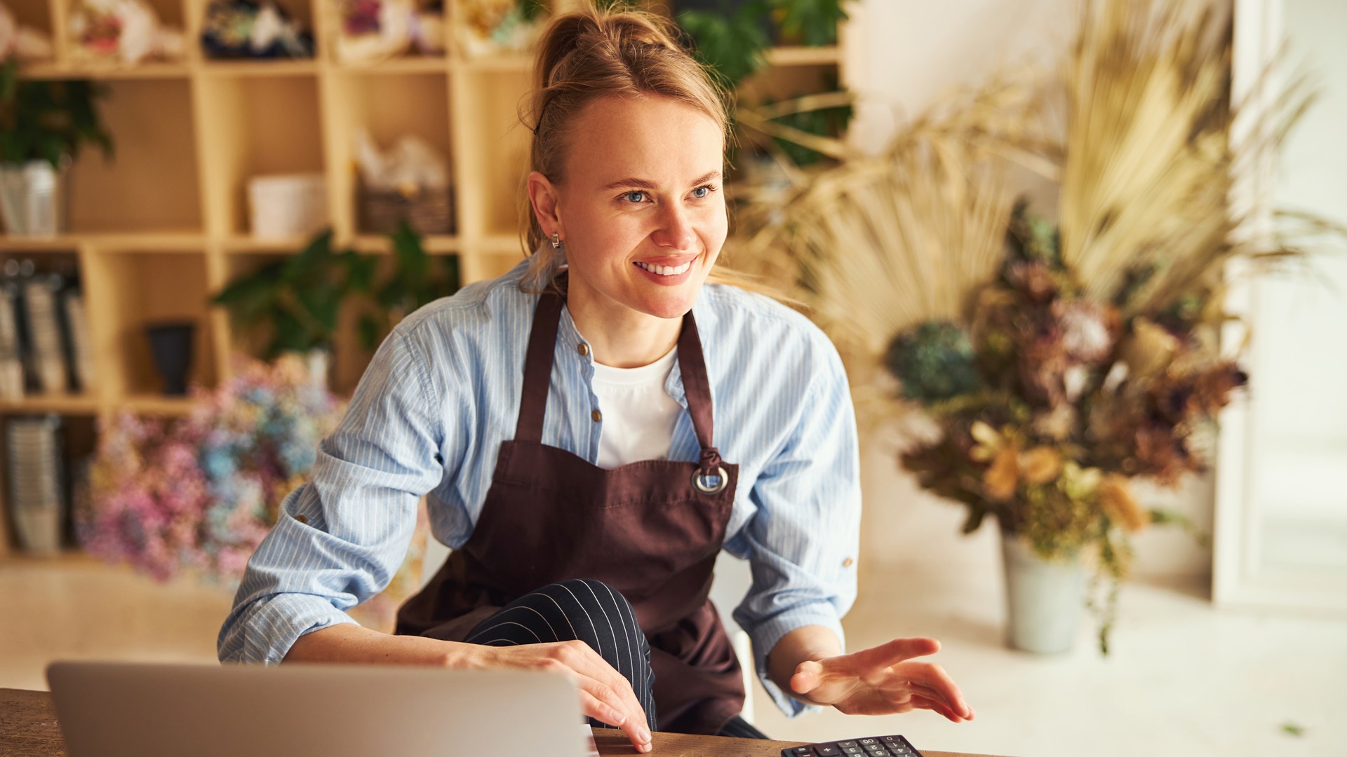 Smiling woman wearing an apron behind a counter with a laptop and plants nearby.