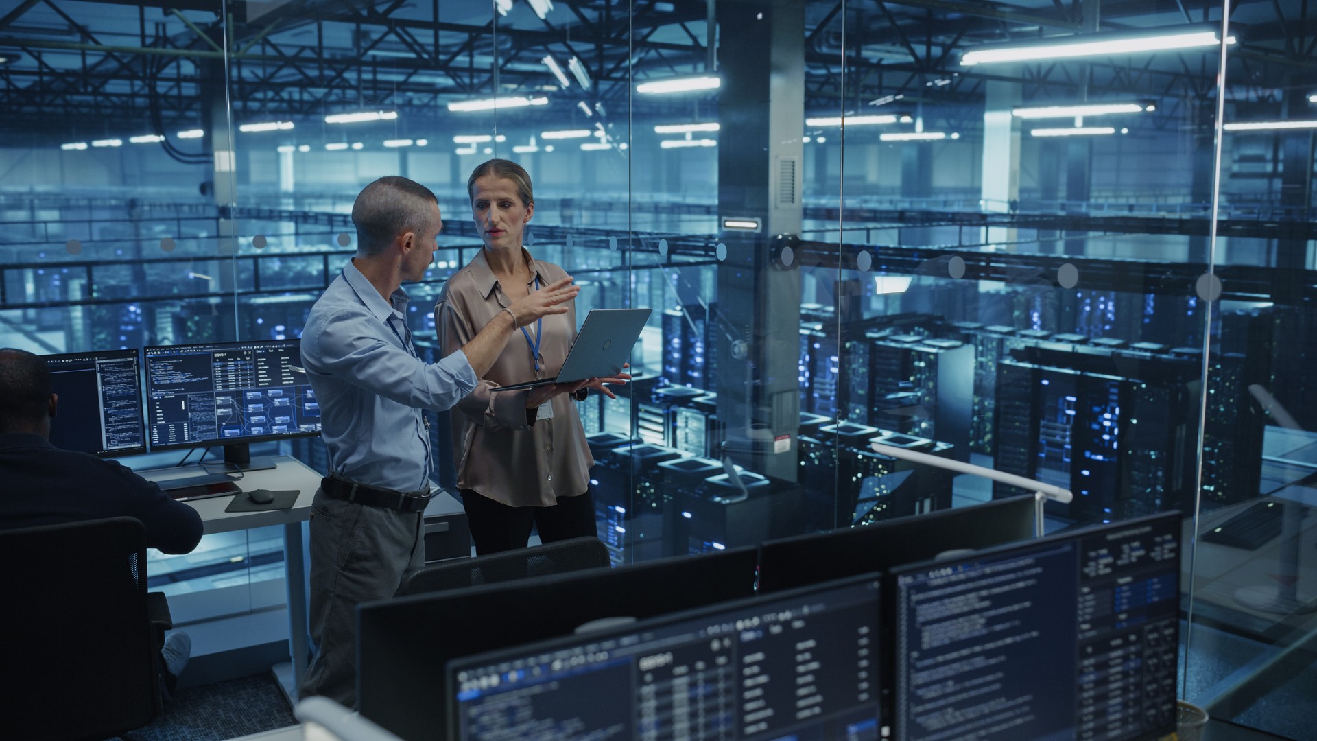 Two people talking with a laptop in a glass-walled area overlooking server equipment.