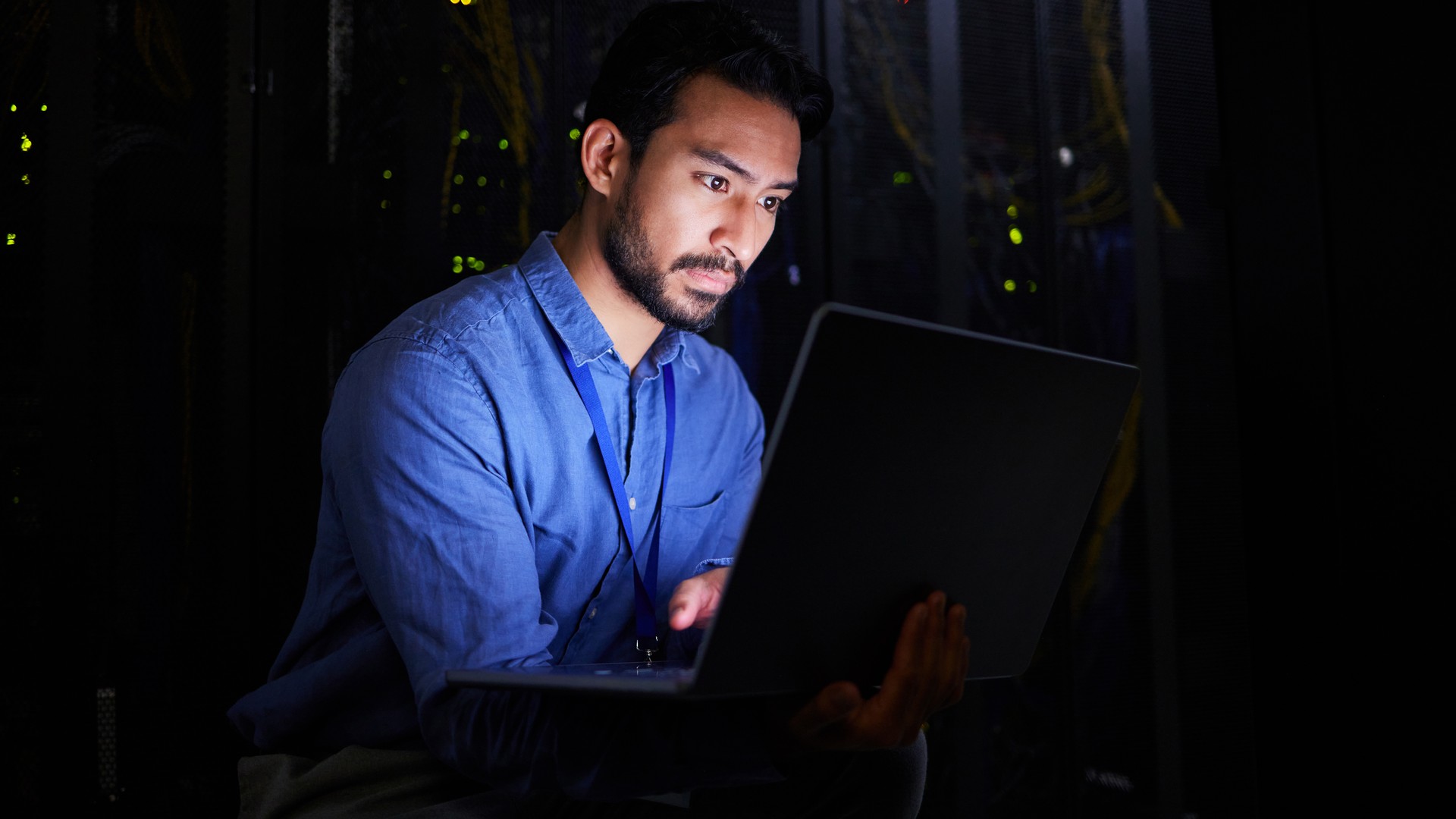 Man using a laptop in a dim room with equipment racks behind him.