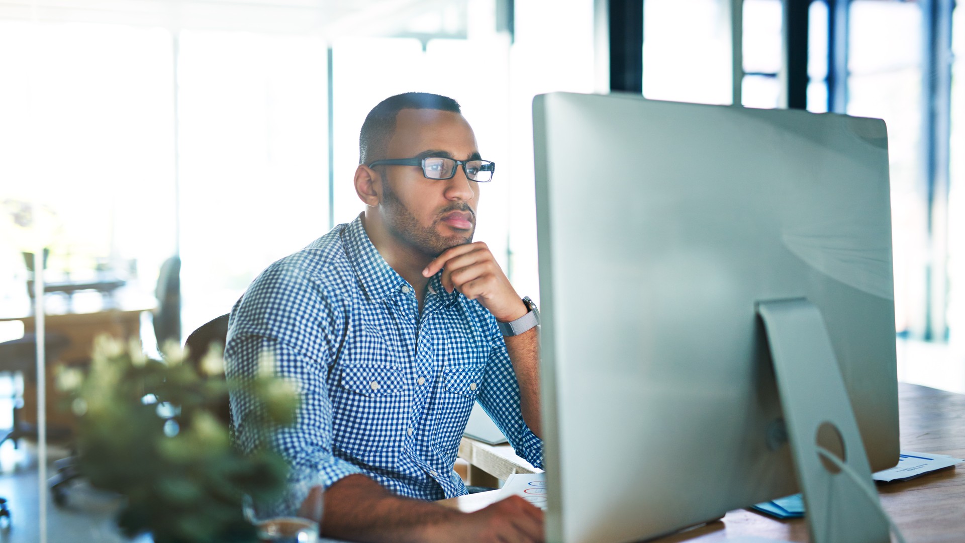 Person wearing glasses looking at a desktop monitor with a hand resting at their chin.