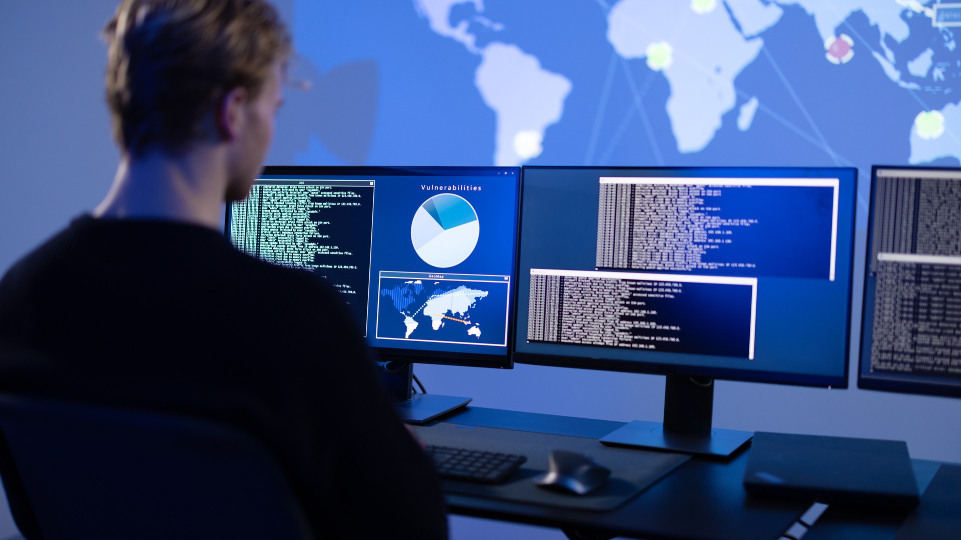 Person faces several computer monitors showing text, maps and a pie chart.
