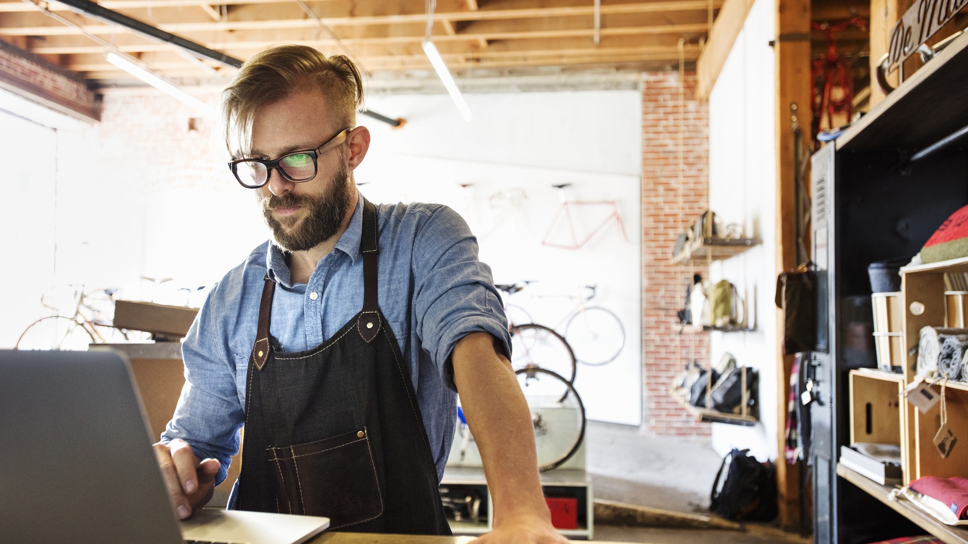 Man wearing glasses and an apron using a laptop at a shop counter.