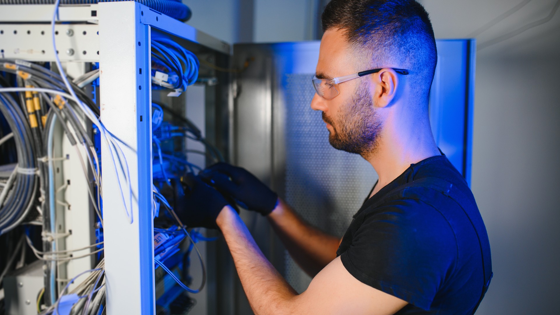 Man wearing glasses and gloves working with cables inside an open equipment cabinet.