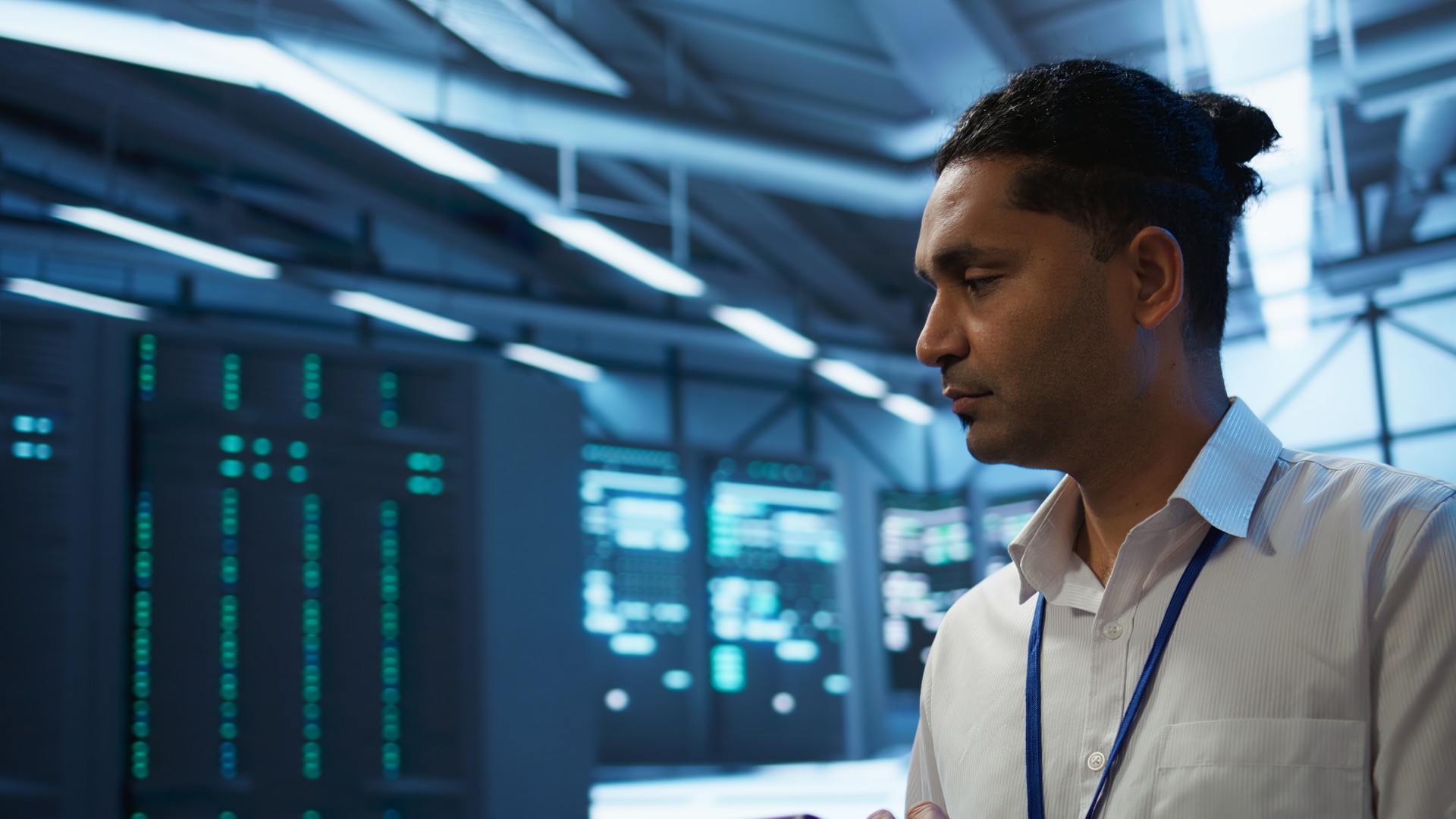 Man wearing a lanyard beside equipment racks in a blue-lit room.
