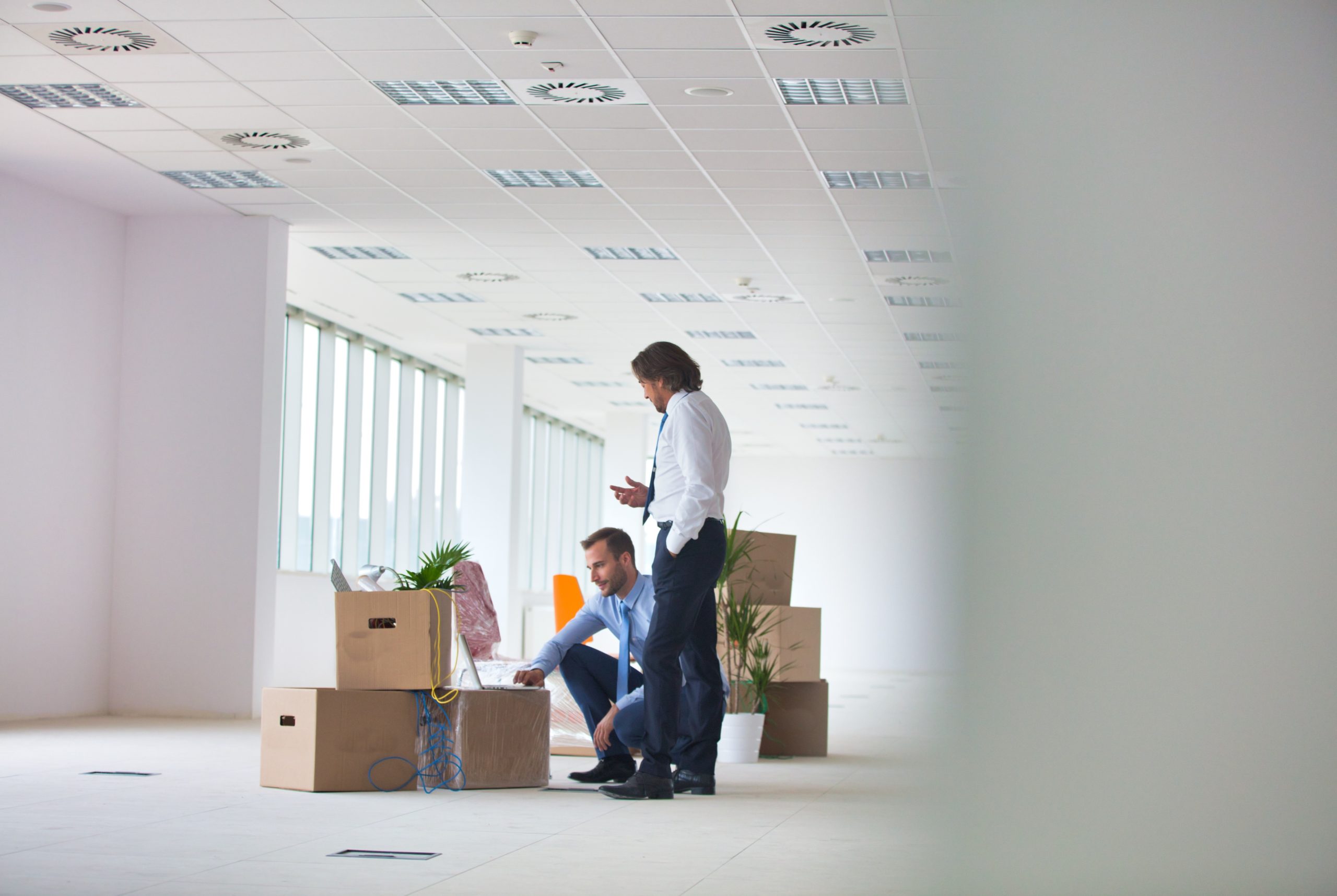 Patch Management image by O&O Systems Two men beside cardboard boxes in a mostly empty office, one crouching at a laptop while the other stands.