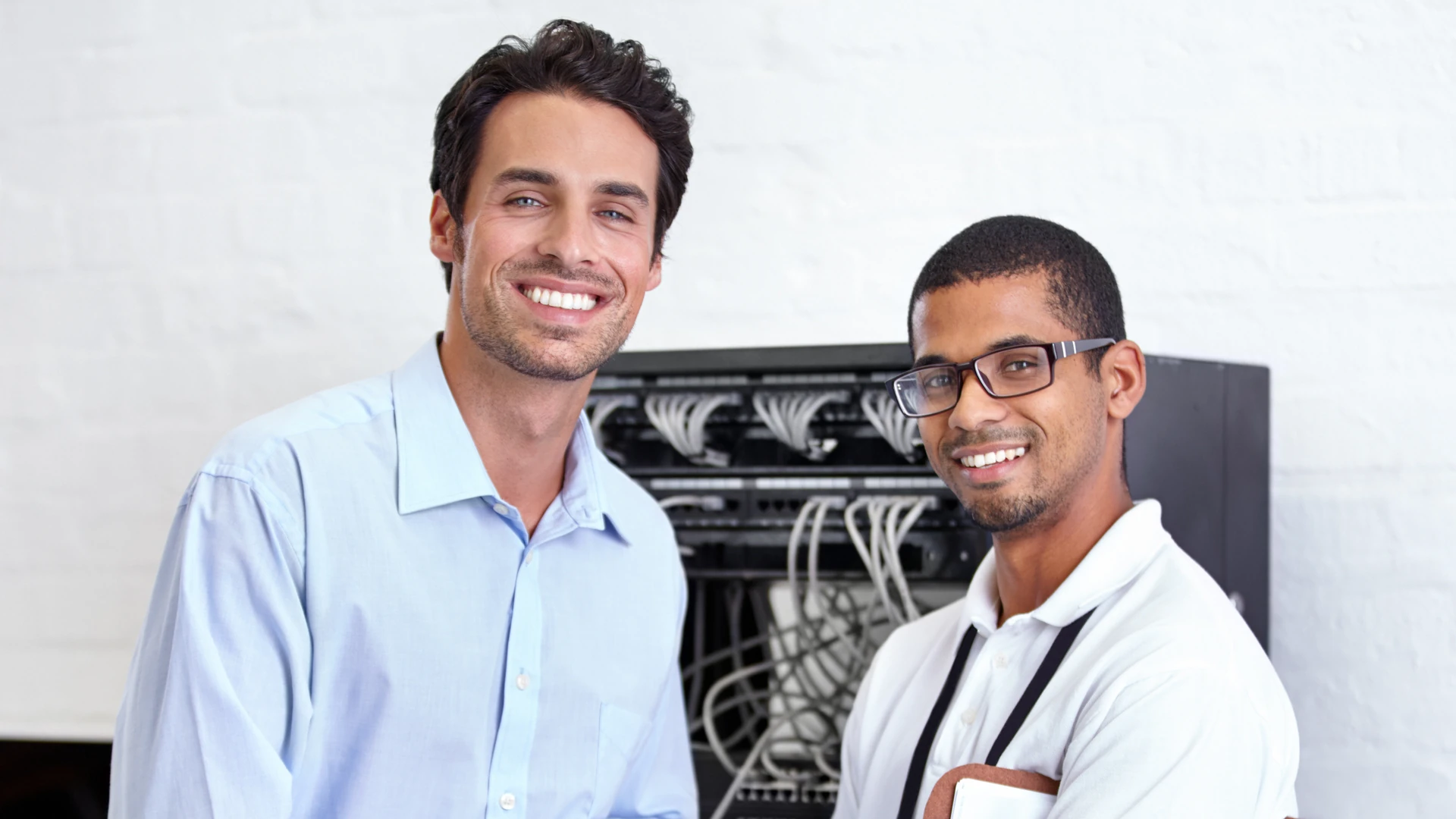 Two smiling men standing in front of an open network rack with patch cables.