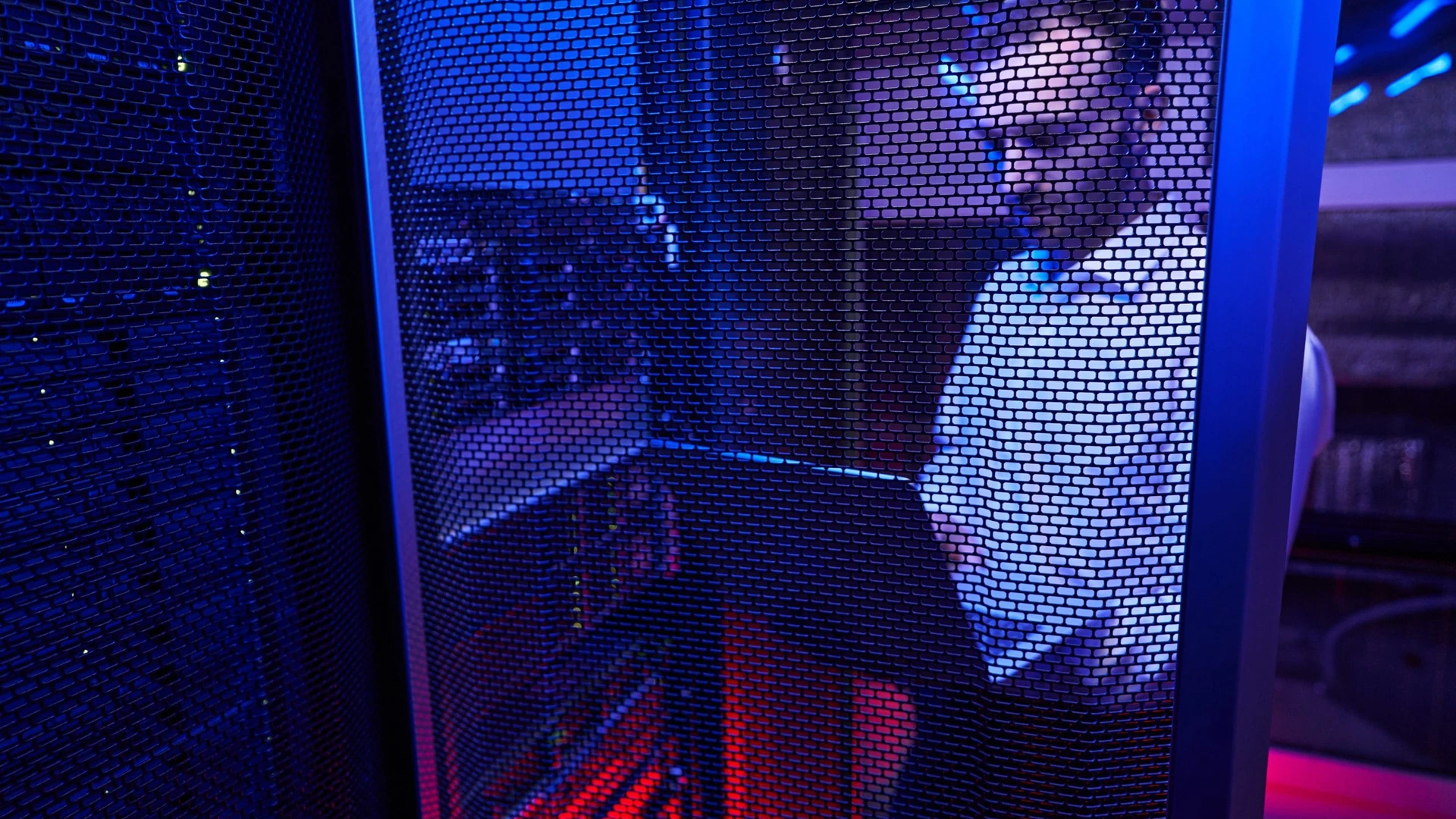 Man looking down at a laptop, seen through the perforated mesh door of a server cabinet in blue and red light
