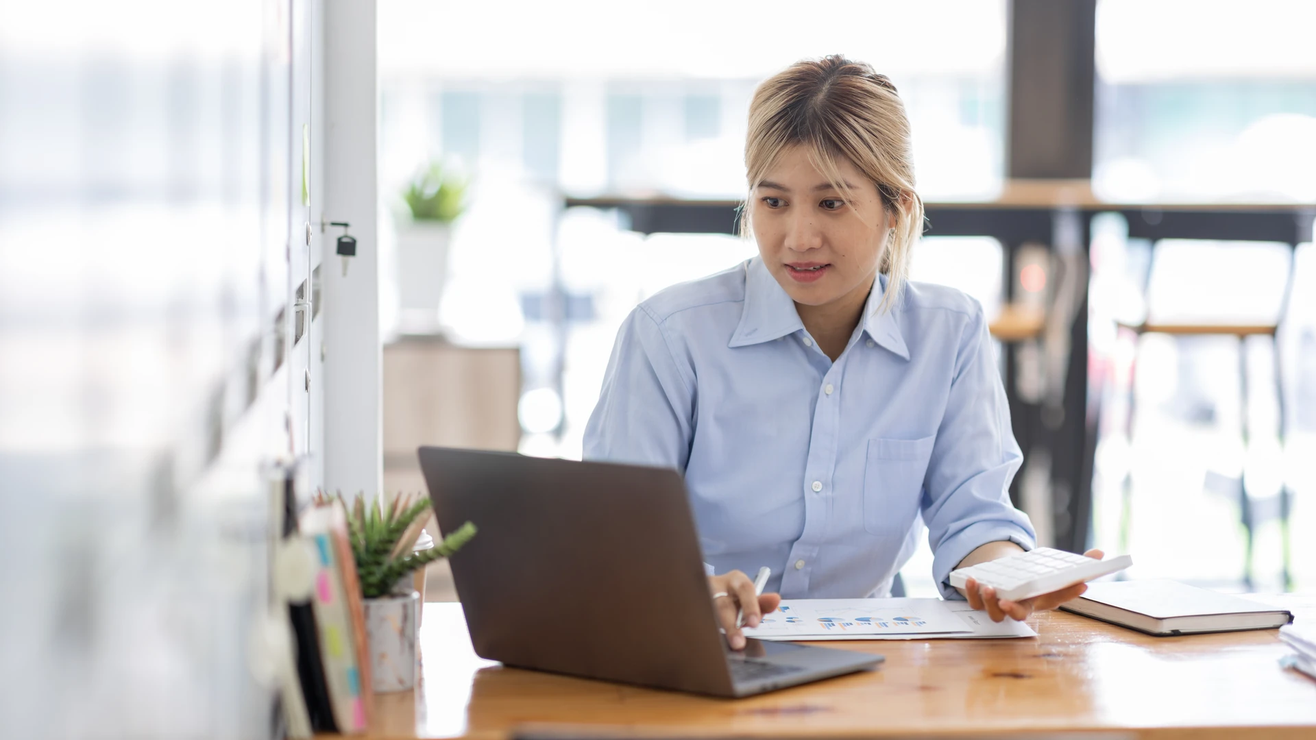 Patch Management image by O&O Systems A woman in a light blue shirt looks at an open laptop at a wooden desk, holding a white calculator, with printed charts on the desk in front of her