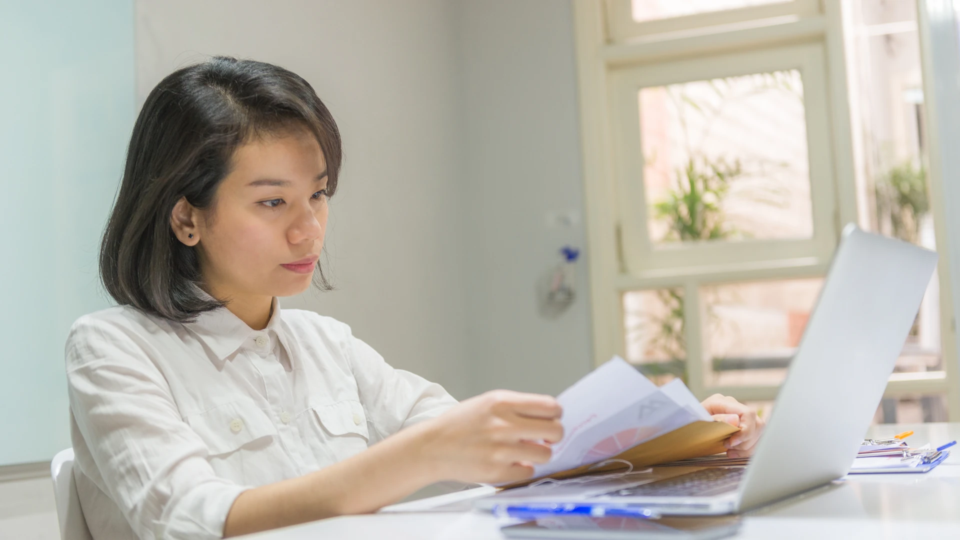 A woman in a white button-up shirt sits at a white desk in a bright office and looks toward an open laptop while holding a few sheets of paper and a tan folder, with a clipboard and pens at the edge of the desk and a window with plants behind her
