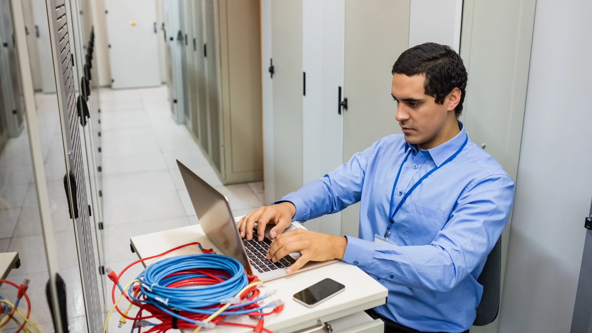 A man in a blue shirt and blue lanyard types on a laptop at a small white desk in a corridor lined with tall gray cabinets, with coiled blue and red network cables and a phone on the desk beside the laptop