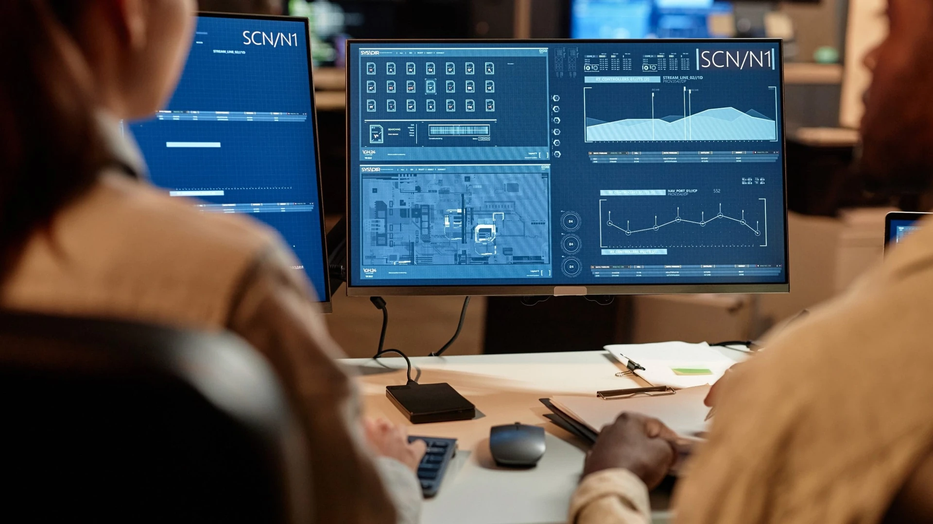 Home image by O&O Systems Two people seen from behind look at a desktop monitor showing charts and status panels on a dark blue screen, with a second monitor beside it and papers on the desk
